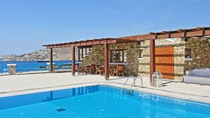 Piscina con pared de piedra y pérgola de madera, con vistas a la costa.