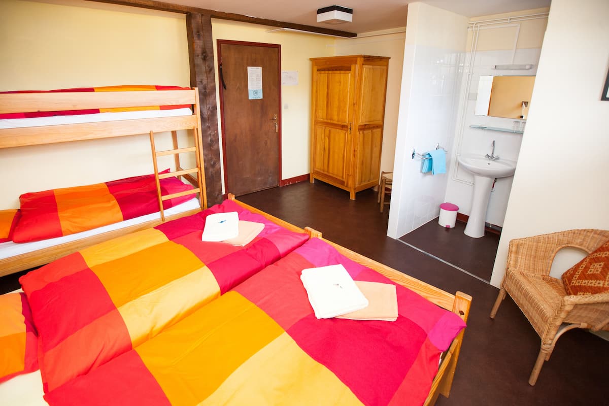 A room with bunk beds, a wooden wardrobe, a bathroom with a sink and mirror, and a wicker chair.