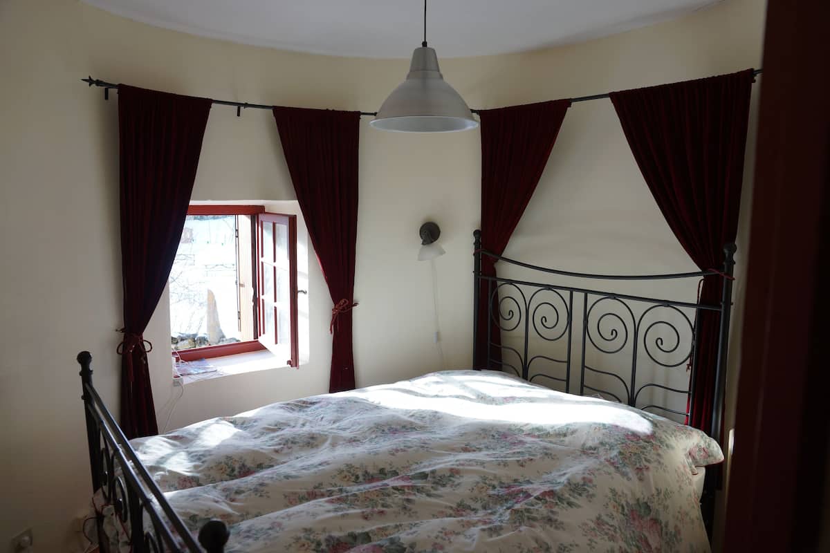 A bedroom with a bed, a window with curtains, a hanging light fixture, and a wall-mounted clock.