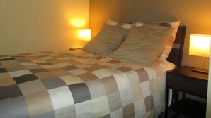 Ein ordentlich bezogenes Bett mit karierter Tagesdecke, zwei Nachttischlampen und einem gerahmten Bild an der Wand.