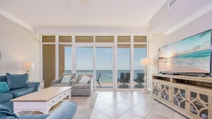 A living room with a large TV, a sofa, a patterned armchair, and a view of the ocean through large glass doors.