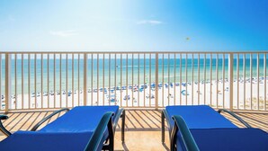 A balcony with two blue lounge chairs overlooking a beach with numerous umbrellas and people.