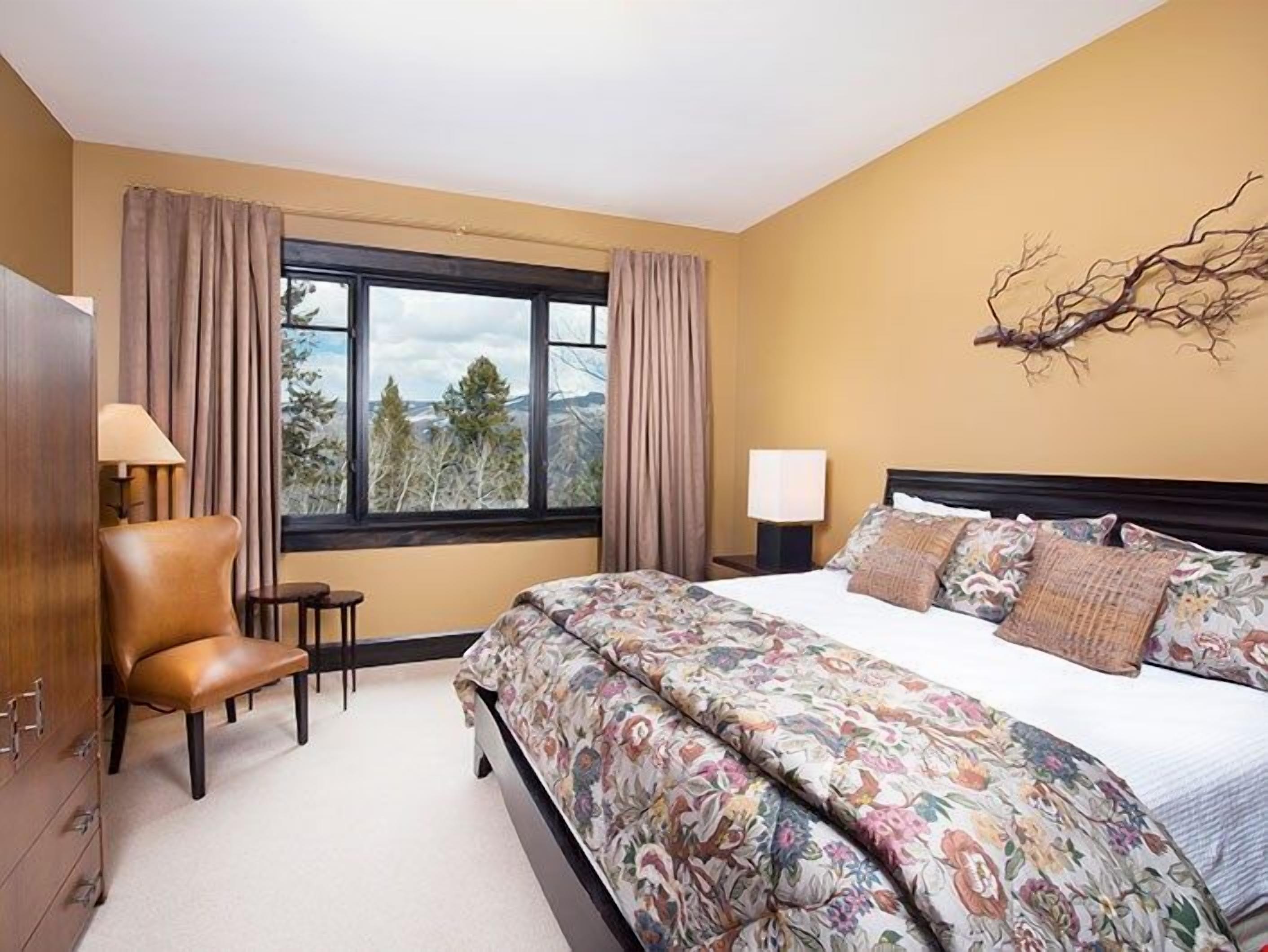 Ein Schlafzimmer mit einem großen Bett, einem Sessel, einem Schreibtisch und einem Fenster mit Vorhängen.