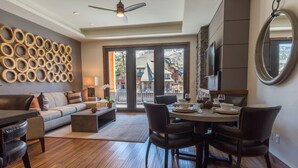 A modern living room with a sofa, coffee table, and dining area with chairs. There is a fireplace, a ceiling fan, and a large window with a view of buildings outside.
