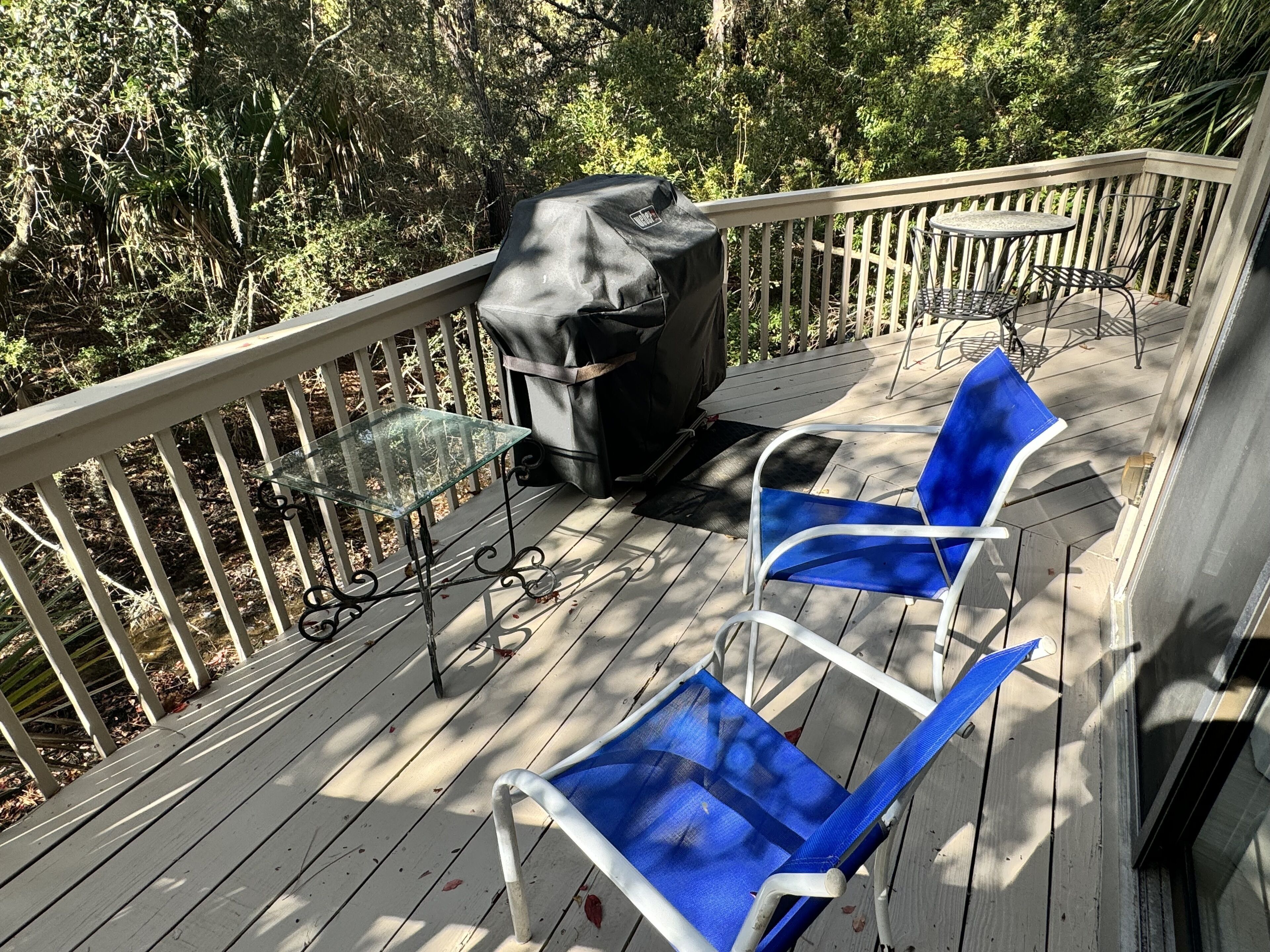 A wooden deck with a glass table and chairs, a barbecue grill, and a view of dense foliage.