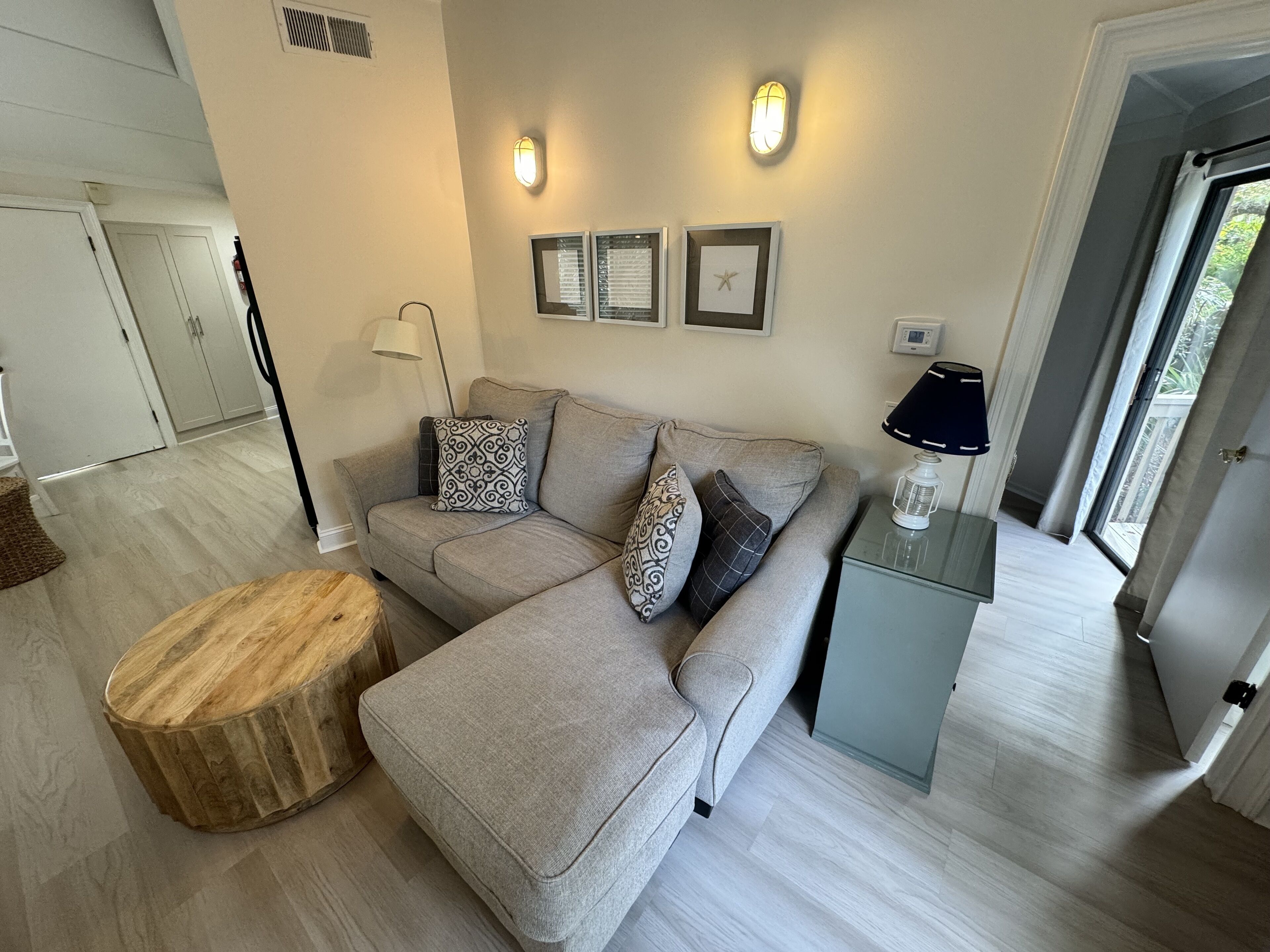 A modern living room with a grey sectional sofa, a wooden ottoman, and a small side table with a lamp. There are framed pictures on the wall and a door leading to another room.