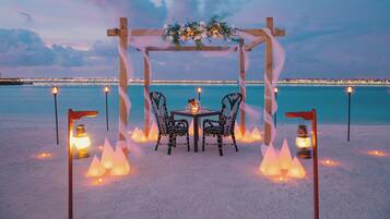 A beachside outdoor dining setup with a wooden arch, two chairs, a table, and decorative lights.