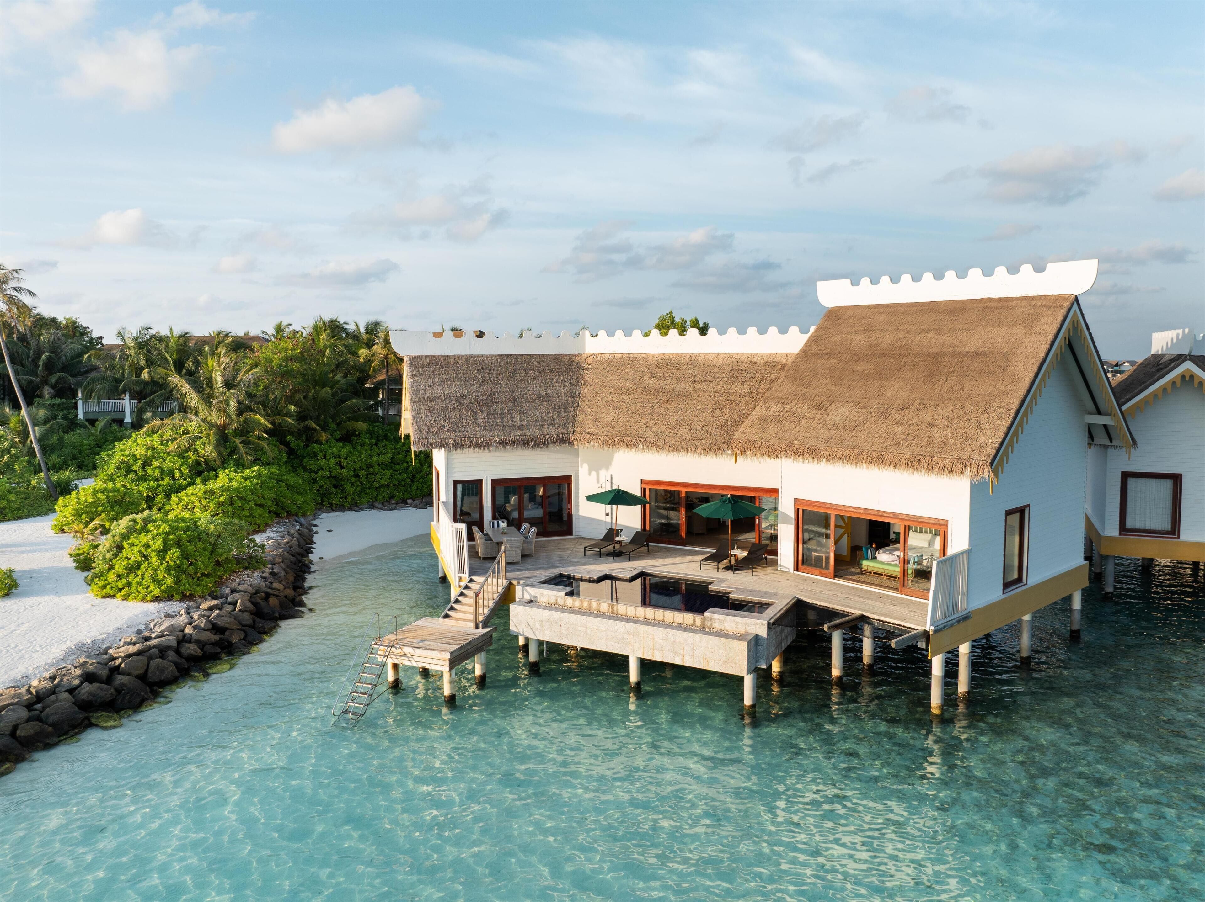 An overwater bungalow with a thatched roof, a pool, and a view of the ocean and palm trees.