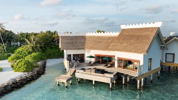 An overwater bungalow with a thatched roof, a pool, and a view of the ocean and palm trees.