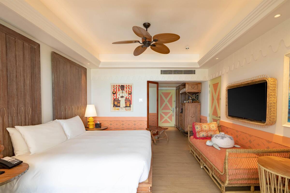 A hotel room with a large bed, a ceiling fan, a TV, a wicker sofa, and a small table.