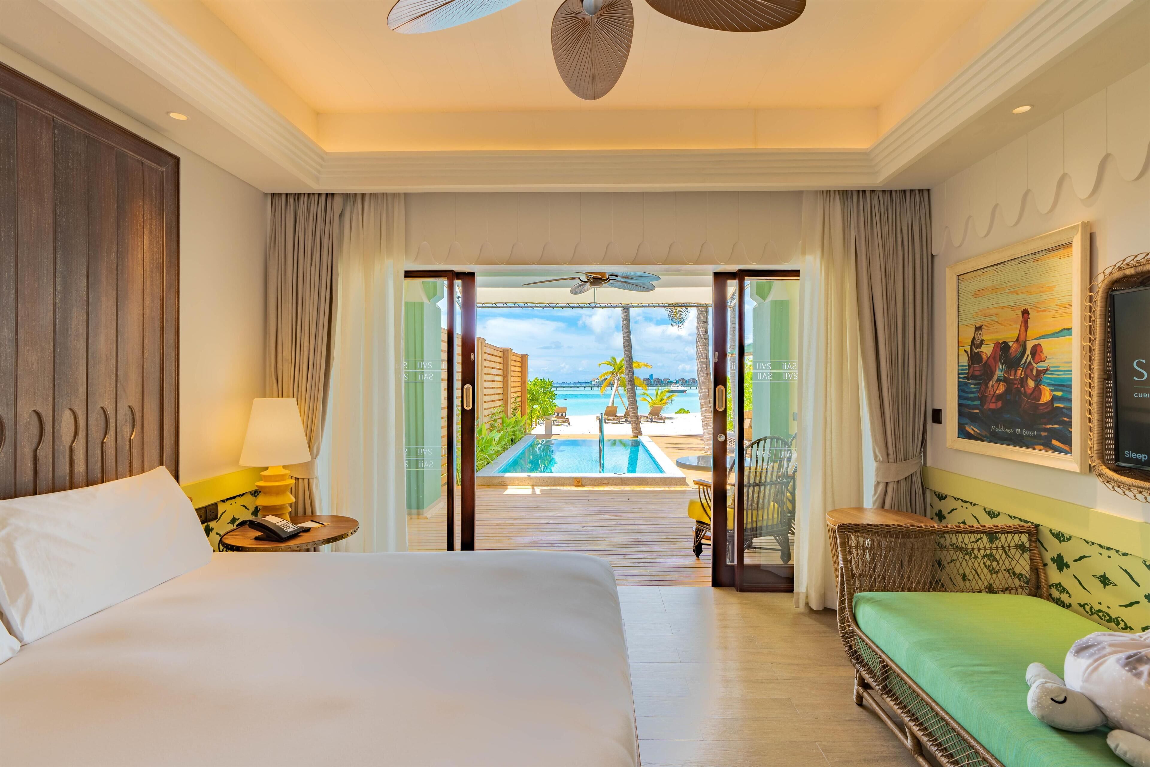 Habitación de hotel con cama, sofá verde, un peluche, lámpara, teléfono y vistas a una piscina y palmeras.