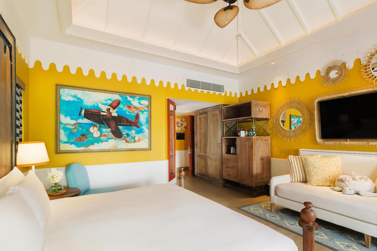 A hotel room with a large bed, a wooden wardrobe, a sofa, a ceiling fan, and a painting of an airplane.