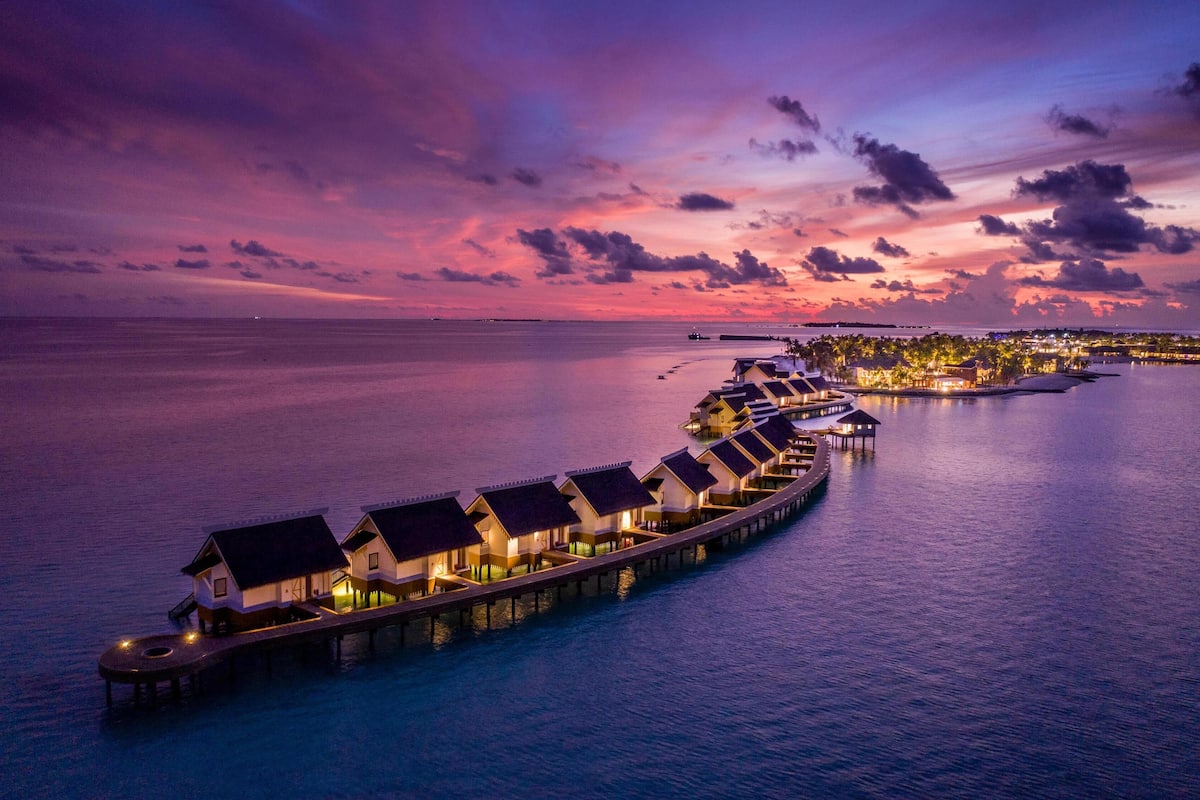 A row of overwater bungalows connected by a pier, with a sunset over the ocean.