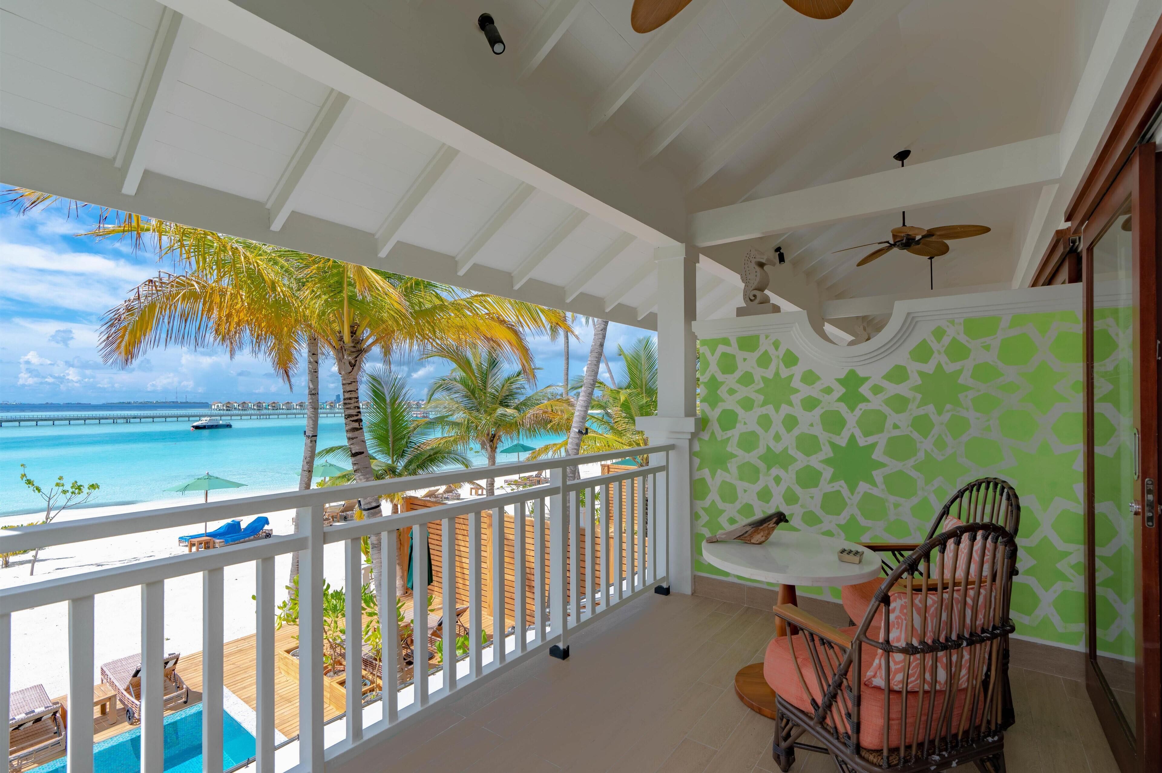 A balcony with a view of a beach and ocean, a dining area with a table and chairs, and a ceiling fan.