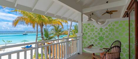 A balcony with a view of a beach and ocean, a dining area with a table and chairs, and a ceiling fan.