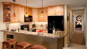 A kitchen with wooden cabinets, granite countertops, a built-in stove, and a refrigerator. There are three bar stools and a wine bottle on the counter. - Condo near Gondola! Free Shuttle (Avon)