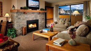 A cozy living room with a stone fireplace, a flat-screen TV mounted above it, and a large window offering a mountain view. - Condo near Gondola! Free Shuttle (Avon)