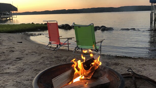 A beach scene with a fire pit in the foreground, two beach chairs, and a calm lake in the background. - Terrific Family Getaway on Lake Charleviox with Stunning Views  (East Jordan)