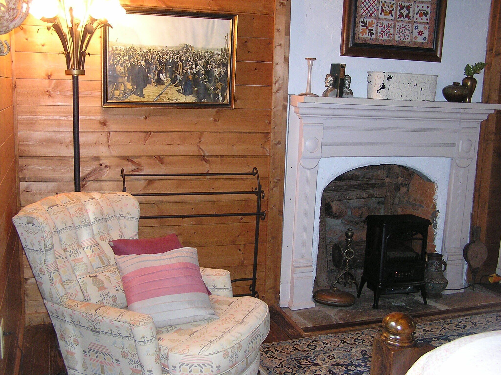Een gezellige kamer met een houten wand, een open haard, een ingelijst historisch schilderij en een fauteuil met bloemenprint.