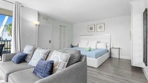 A modern hotel room with a grey sofa, a bed with blue bedding, and a balcony with a view of palm trees. - Beach & Tennis Getaway 3206 (Bonita Springs)