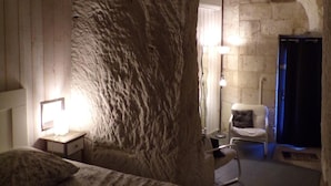 Un dormitorio con pared de piedra, una cama, una silla y una mesita de noche con una lámpara.