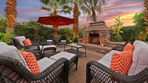 An outdoor patio with wicker furniture, a stone fireplace, and a red umbrella. - Experience luxury at Paramount, featuring stunning mountain views, a large pool and game room! (Indio)