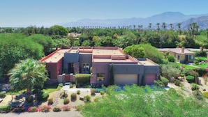 A modern two-story house with a flat roof, surrounded by lush greenery and palm trees. - Experience luxury at Paramount, featuring stunning mountain views, a large pool and game room! (Indio)