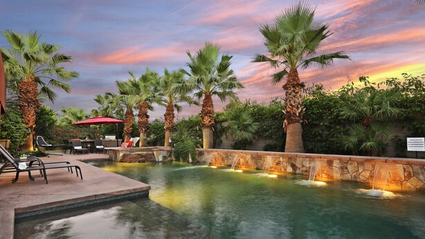A pool area with palm trees, a stone wall, and a waterfall feature. - Experience luxury at Paramount, featuring stunning mountain views, a large pool and game room! (Indio)
