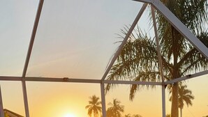 Un espacio de estar techado al aire libre con dos sillas azules y un banco, con vistas a palmeras y un atardecer.