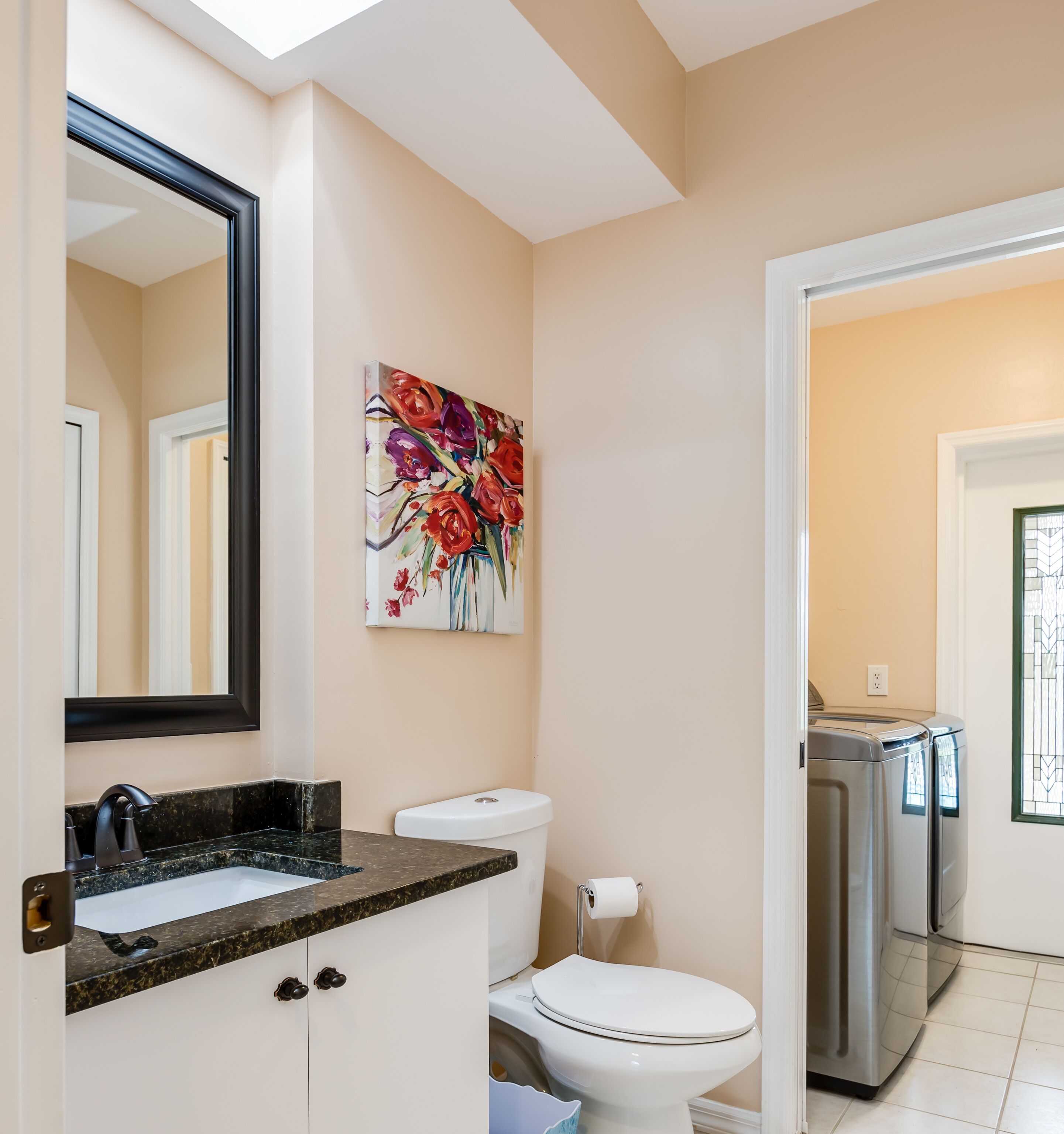 A modern bathroom with a large mirror, a sink with a granite countertop, a toilet, and a stainless steel washing machine.
