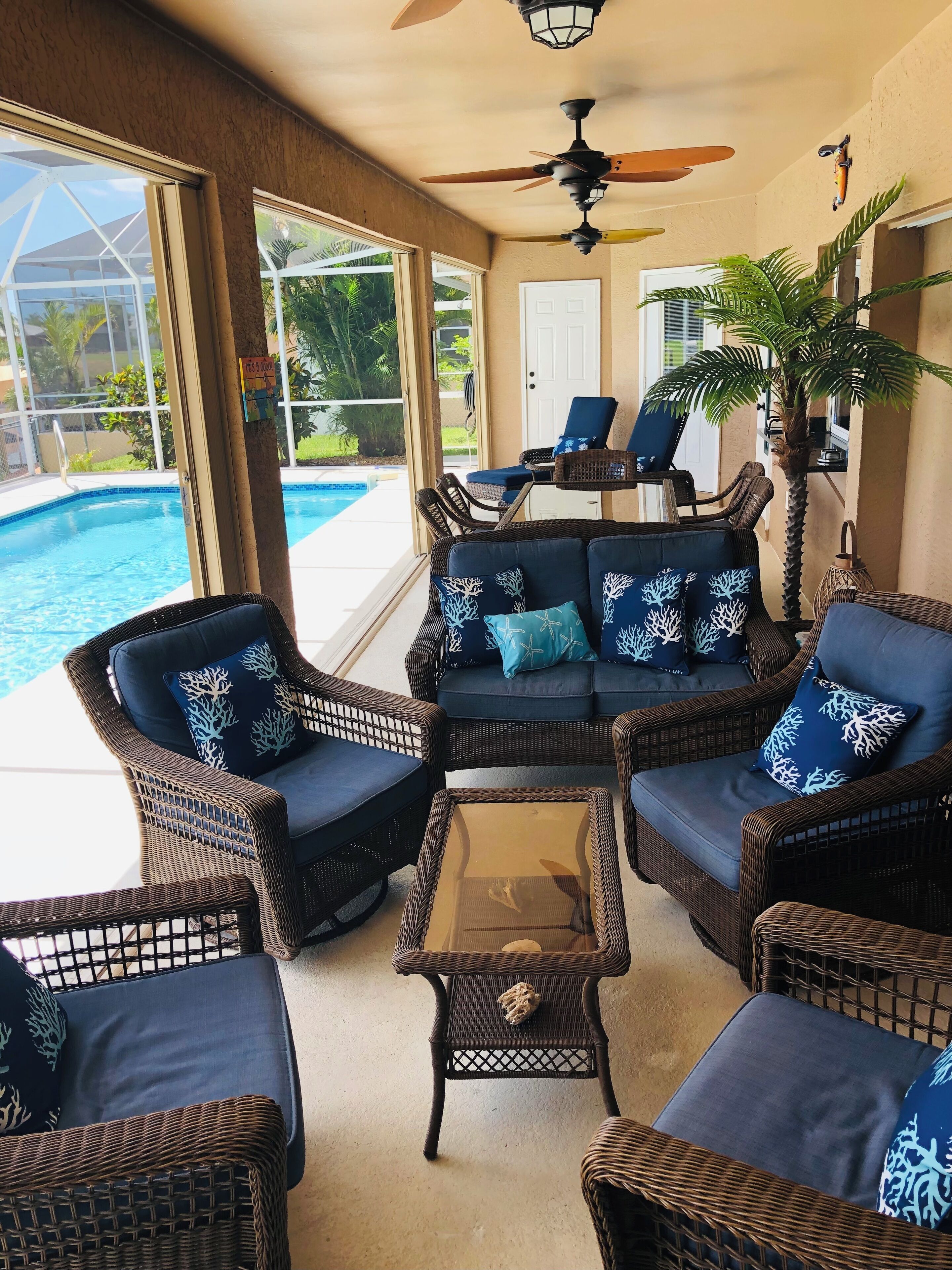 Ein helles Sitzareal im Innenbereich mit Korbmöbeln, Blick auf einen Pool und Deckenventilatoren.