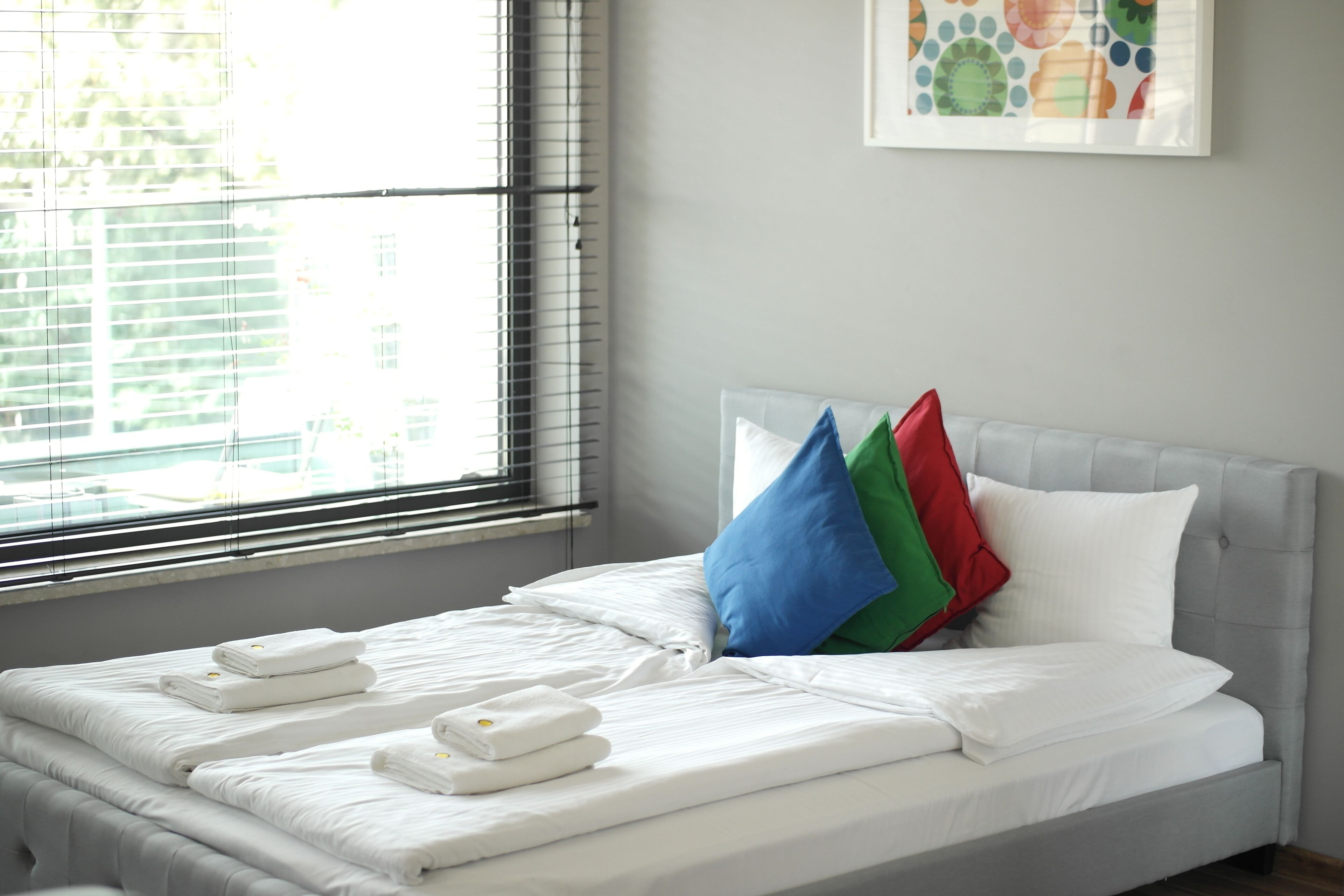 A neatly made bed with white linens and towels, a grey headboard, and a colorful wall art piece above it.