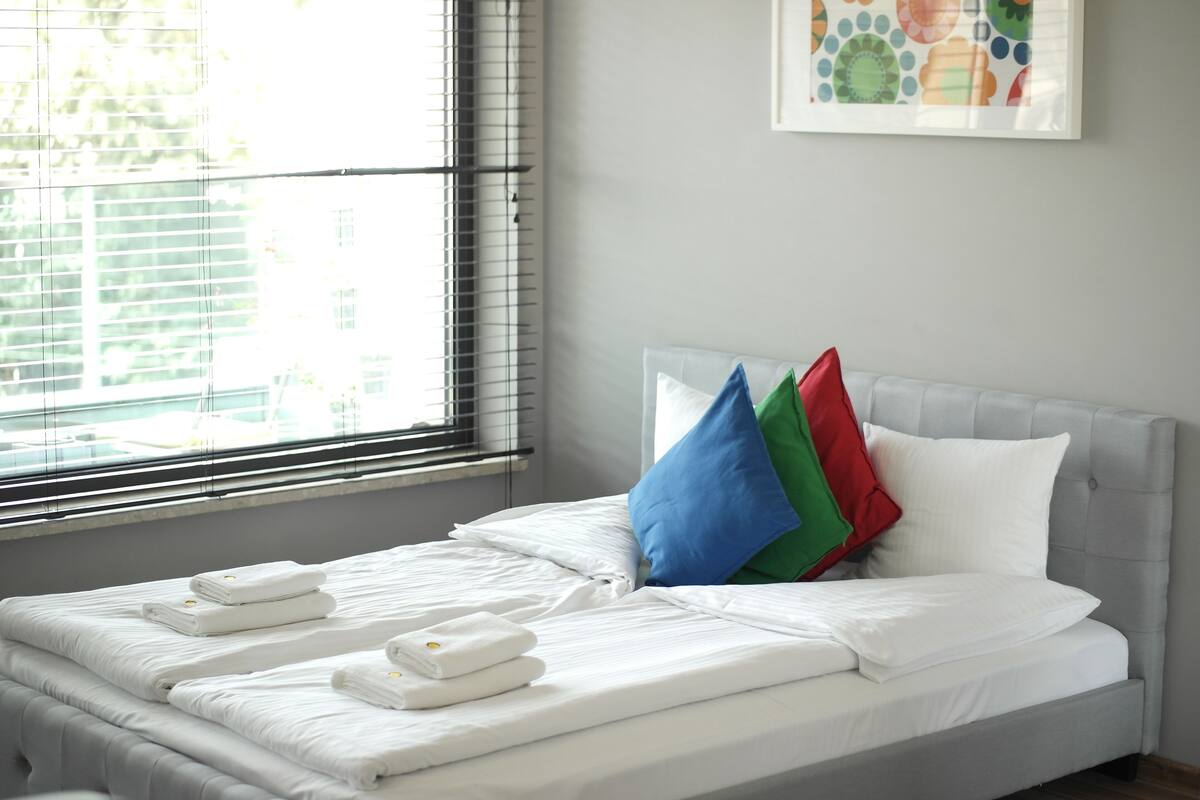 A neatly made bed with white linens and towels, a grey headboard, and a colorful wall art piece above it.