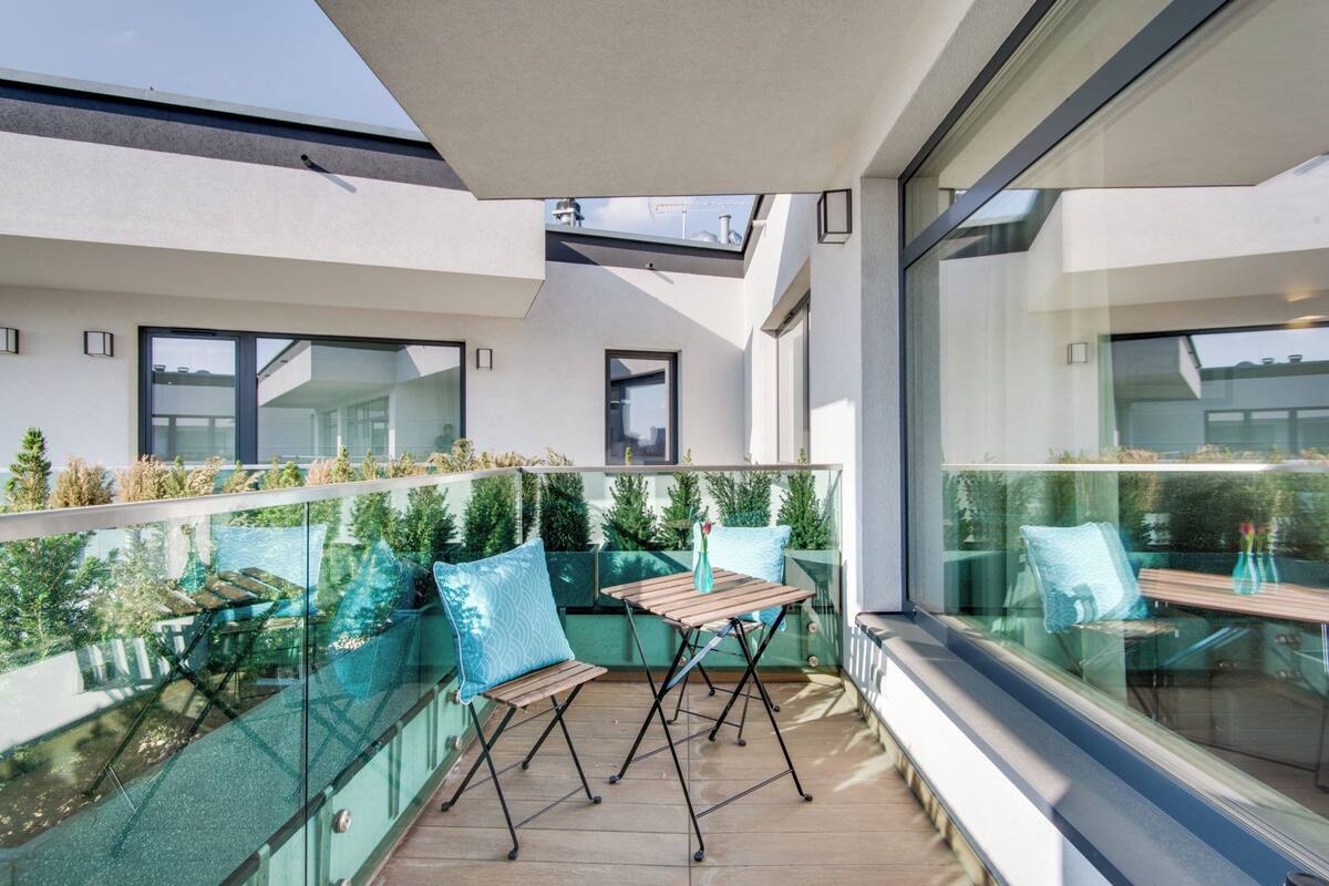 A modern balcony with glass railings, wooden furniture, and potted plants.