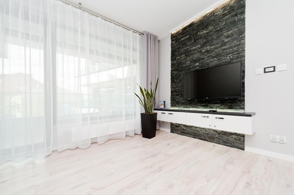 A modern living room with a large flat-screen TV mounted on a stone wall, white cabinetry below, and a large window with sheer curtains.