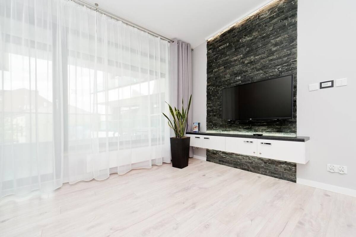 A modern living room with a large flat-screen TV mounted on a stone wall, white cabinetry below, and a large window with sheer curtains.