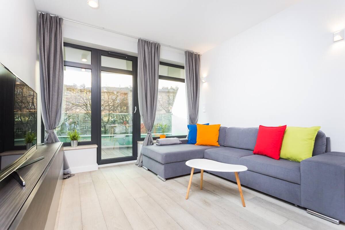A modern living room with a grey sofa, colorful cushions, a white coffee table, and a large window with curtains.