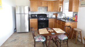 A compact kitchen with wooden cabinets, a stainless steel refrigerator, a microwave, a stove, and a sink. There is a dining table with chairs and a small stool. - Ocean View And Breezes, Quiet 1 Bedroom, 1 Bath Private Guest Cottage On 1 Acre (Christiansted)