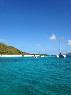 A clear blue sea with several sailboats anchored near a sandy beach with white sand and green hills in the background.