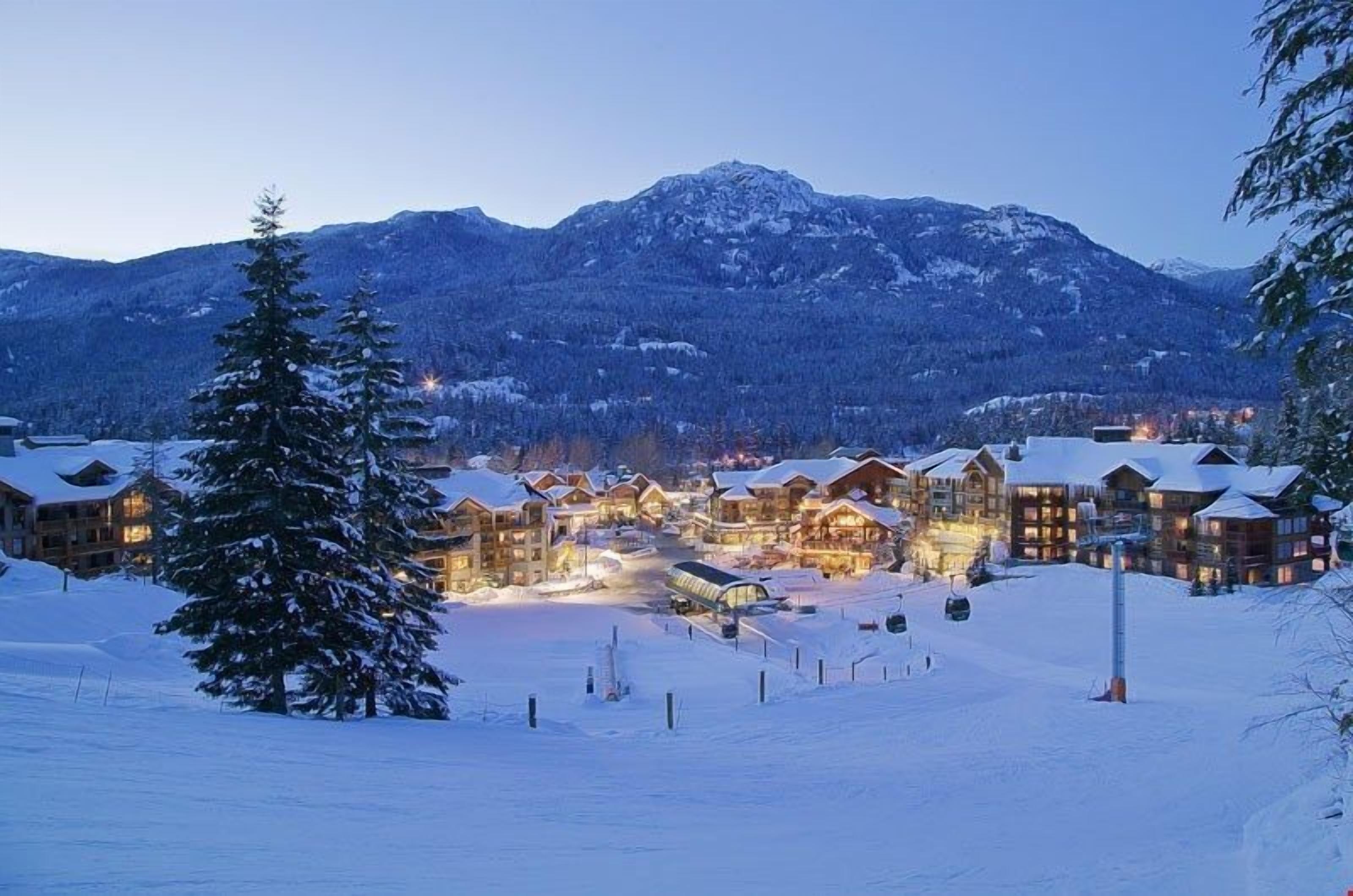 A snow-covered lodge with multiple buildings, a ski lift, and a mountain backdrop.