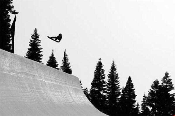 Um snowboarder no ar, executando um salto em uma encosta nevada, com altas árvores perenes ao fundo.