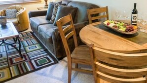 Ein gemütliches Wohnzimmer mit einem dunkelbraunen Ledersofa, Holzstühlen, einem runden Holztisch mit Obstplatte und Weingläsern sowie einem farbenfrohen Teppich.