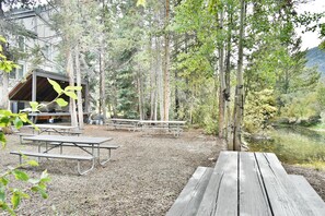 A rustic outdoor dining area with picnic tables and a covered shelter, surrounded by trees and a body of water. - Pet Friendly On River! Pool, Hot Tubs! Walk to Main St. Shops, Restaurants (Frisco)