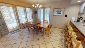 A dining area with a wooden table and chairs, large windows with blinds, a chandelier, and a kitchen counter with utensils. - Pet Friendly On River! Pool, Hot Tubs! Walk to Main St. Shops, Restaurants (Frisco)