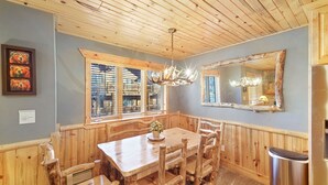 A wooden-paneled dining area with a table and chairs, a chandelier, a window with blinds, and a mirror on the wall.