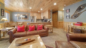 A cozy living room with a wooden ceiling, a leather sofa with red cushions, a wooden coffee table, and a kitchen area with wooden cabinets and a refrigerator.