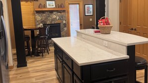 A kitchen with a large island, wooden flooring, stone fireplace, and a dining area with chairs.