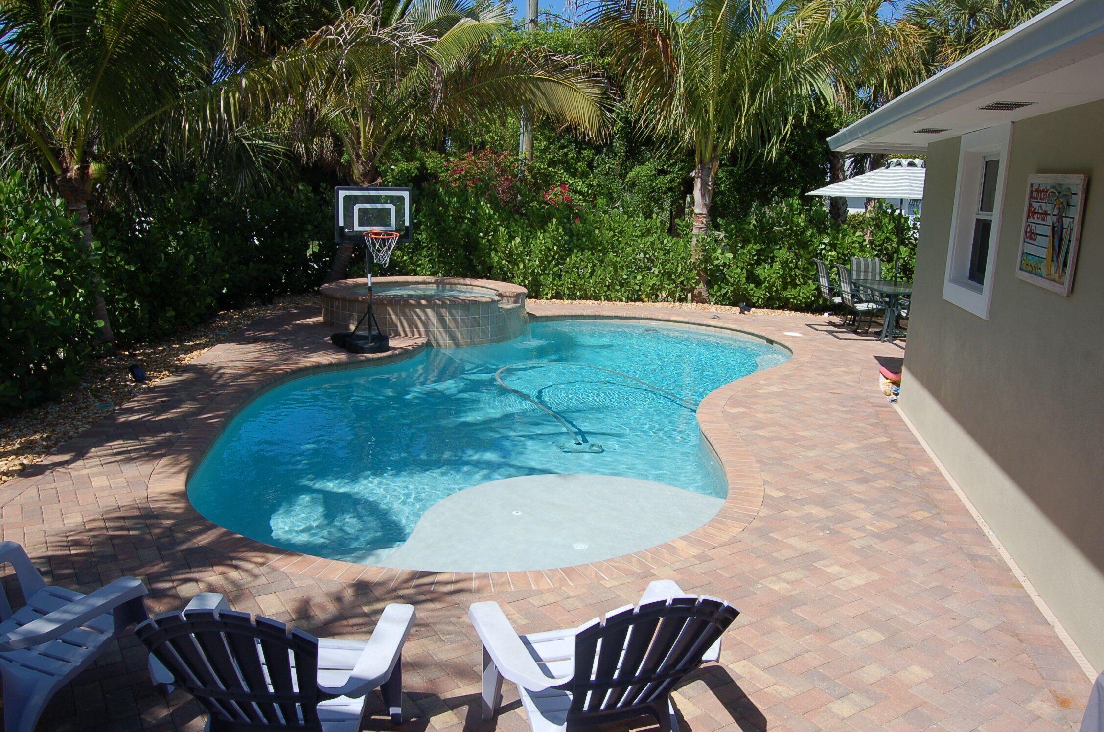 Une piscine avec un panier de basket, entourée de palmiers et dotée d’un espace patio avec des chaises.