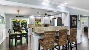 A modern kitchen with white cabinets, a central island with bar stools, and a dining area with a wooden table and chairs. - Well Appointed Home Salt Water Pool & Separate Pool House & Putting Green (Hobe Sound)