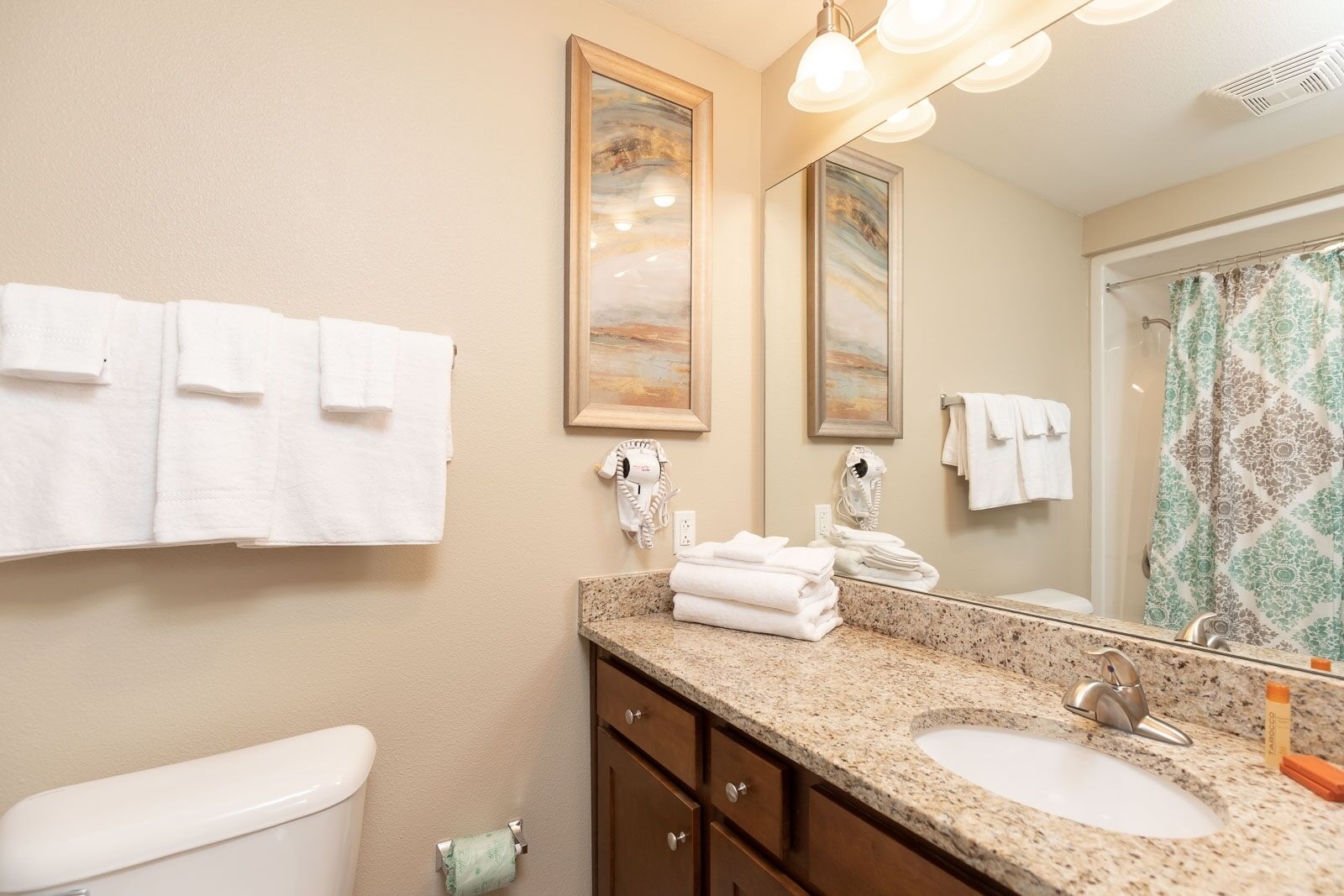 A bathroom with a granite countertop, a sink, a toilet, and a shower with a patterned curtain. There are towels hanging on the wall and a hairdryer on the counter.